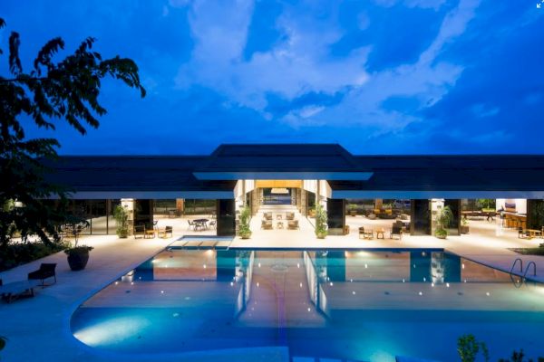 The image shows a modern building with a large swimming pool in the foreground, sparkling under a twilight sky.