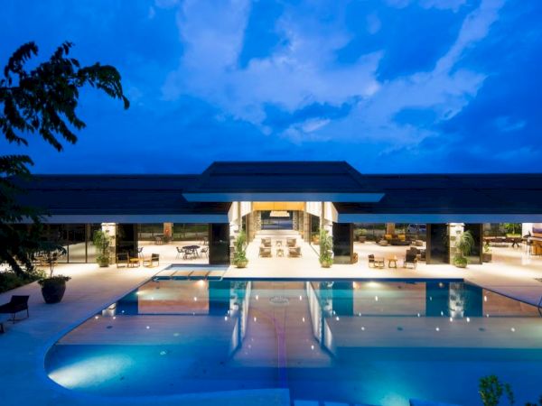 The image shows a modern building with a large swimming pool in the foreground, sparkling under a twilight sky.