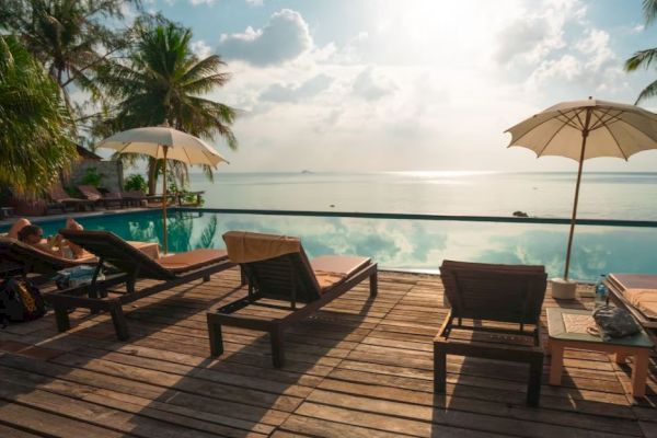 A scenic poolside view with lounge chairs, umbrellas, and palm trees overlooking a tranquil ocean under a partly cloudy sky.