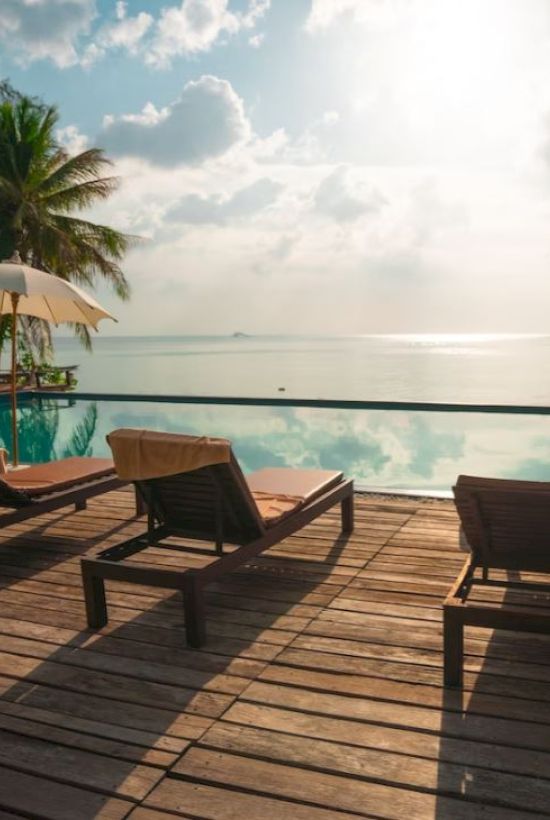 A serene beachside scene showing lounge chairs and umbrellas on a wooden deck overlooking an infinity pool with a beautiful ocean view, and palm trees.