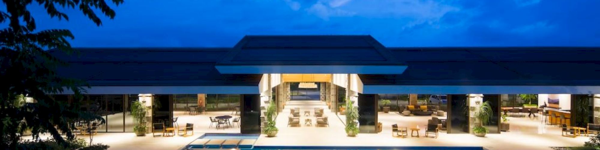 An illuminated resort with a large swimming pool, surrounded by seating areas, under a twilight sky.