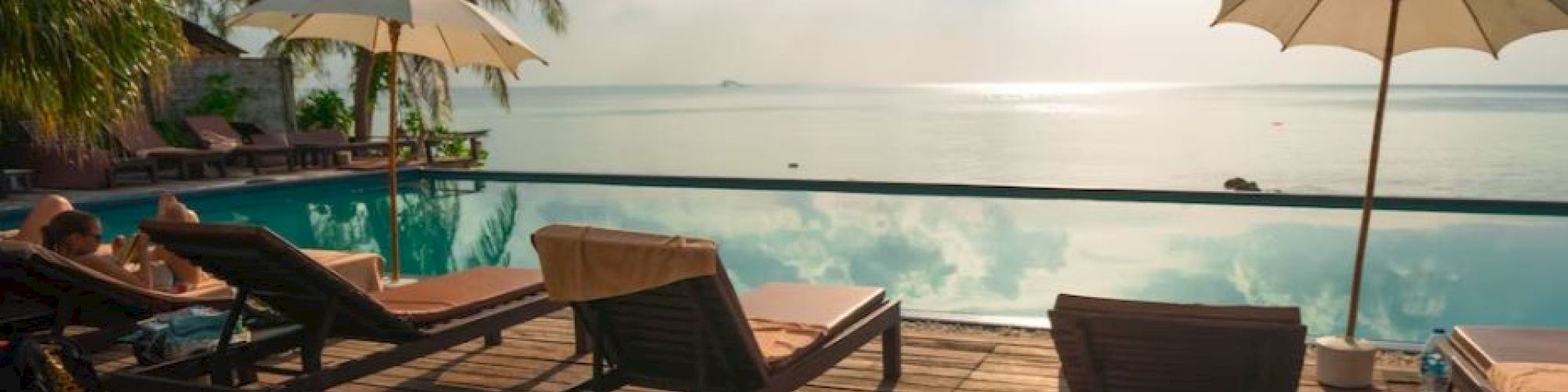 Poolside lounge area with sunbeds and umbrellas overlooking a calm ocean, surrounded by palm trees under a partly cloudy sky.