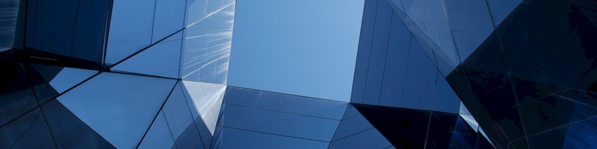 The image shows a view looking up from the center of a modern glass building, depicting reflective surfaces and geometric patterns against a clear sky.
