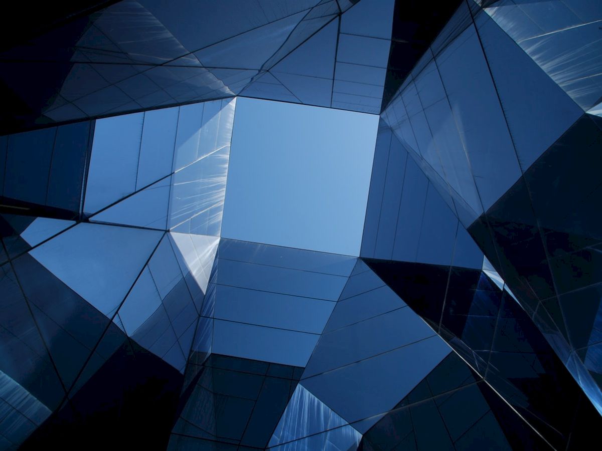 The image shows an upward view of a sharply angular, modern architectural structure with reflective surfaces and a clear sky above.