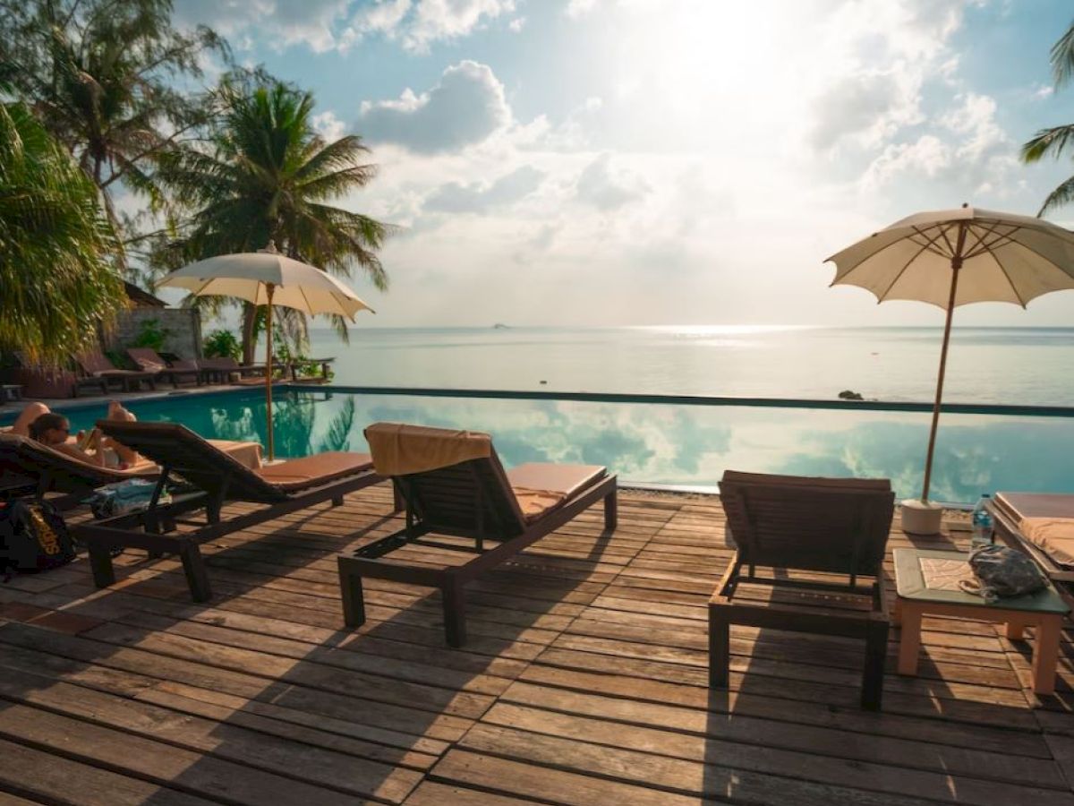 A serene poolside scene with lounge chairs, umbrellas, and lush palm trees overlooking a calm ocean under a partly cloudy sky.