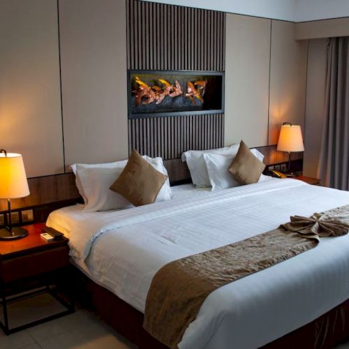 A neatly made double bed in a modern hotel room, flanked by two nightstands with lamps. A wall-mounted fireplace adds a cozy touch.