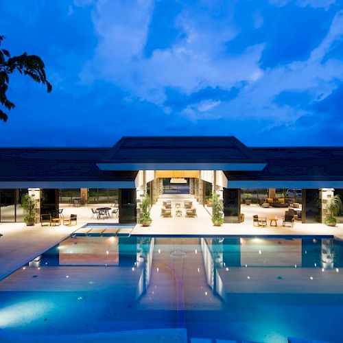 An illuminated modern building features an inviting swimming pool and seating area, set against a picturesque evening sky.