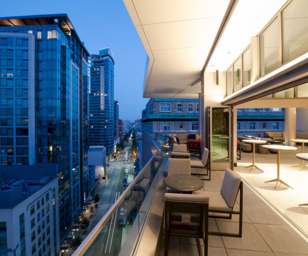A modern rooftop balcony overlooking a cityscape with high-rise buildings. The balcony is furnished with tables and chairs, illuminated at dusk.
