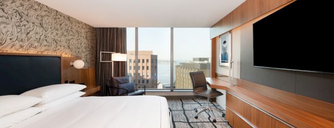 A modern hotel room with a large bed, desk, chair, and a wide window offering a city view, featuring contemporary furniture and decor.