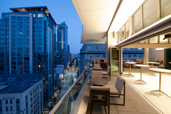 A modern cityscape at dusk with a view from a sleek balcony featuring tables and chairs overlooking high-rise buildings and a street below.