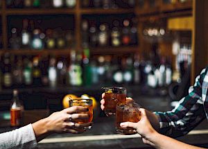 Three people are toasting with drinks in a dimly lit bar, with shelves of bottles visible in the background.