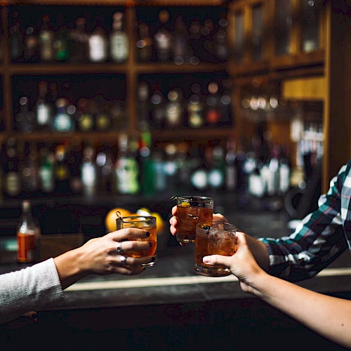 Three people are toasting with drinks in a dimly lit bar, with shelves of bottles visible in the background.