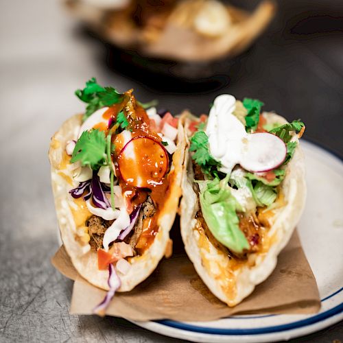 The image shows two loaded tacos on a plate, garnished with various ingredients including lettuce, radish slices, and a creamy sauce.