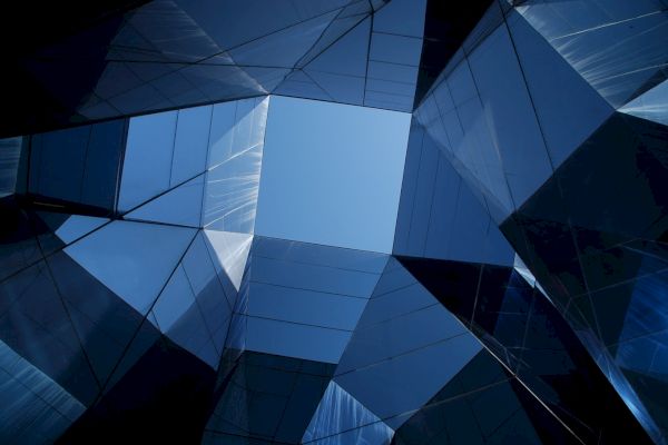 The image shows an upward view of a geometric structure with reflective glass panels, revealing a clear blue sky at the center.