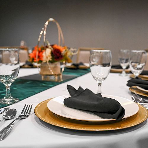 A formal table setting with plates, cutlery, wine glasses, and black napkins on a white tablecloth, featuring a floral centerpiece in a basket.