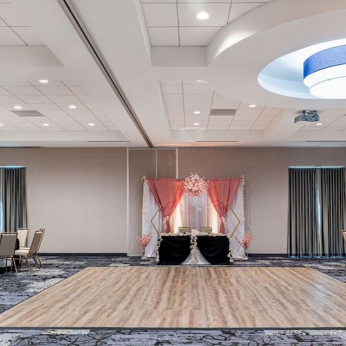 The image shows a decorated event hall with a dance floor, tables, chairs, ceiling lights, and a draped backdrop. It looks ready for a celebration.