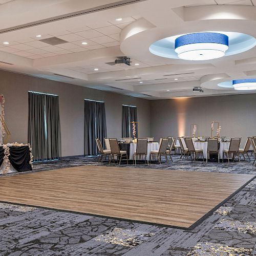 A large, elegantly decorated banquet hall with a wooden dance floor in the center, round tables with chairs, and soft lighting throughout the room.