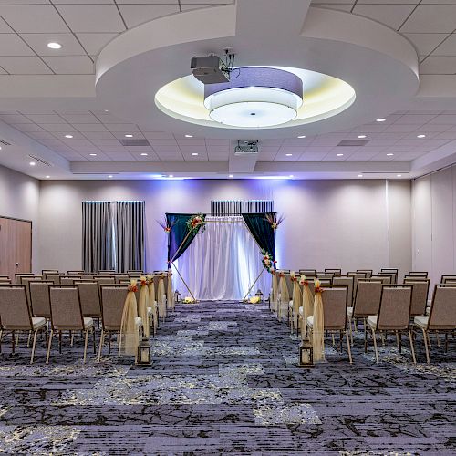 The image shows a decorated indoor venue setup for a formal event with rows of chairs facing a stage with drapery and floral arrangements.