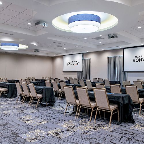 A conference room with rows of chairs and tables facing two screens displaying 