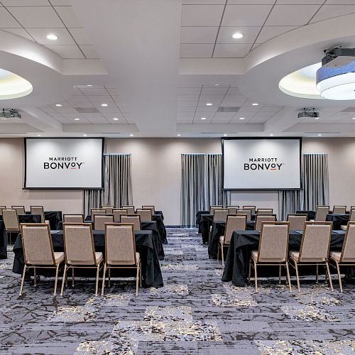 An empty conference room is set up with rows of chairs, two projection screens displaying 