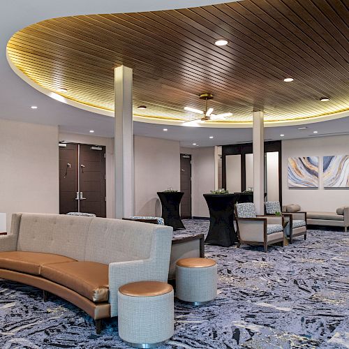 A modern lounge area with beige couches, round tables, wall art, and a circular mirror under a unique wood-paneled ceiling with ambient lighting.