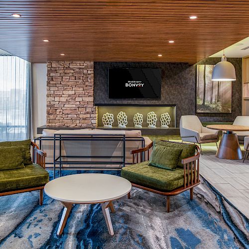 A modern lounge area featuring green chairs, a white round table, a wood-paneled ceiling, a stone accent wall, and a mounted TV displaying “BRAVERY.”