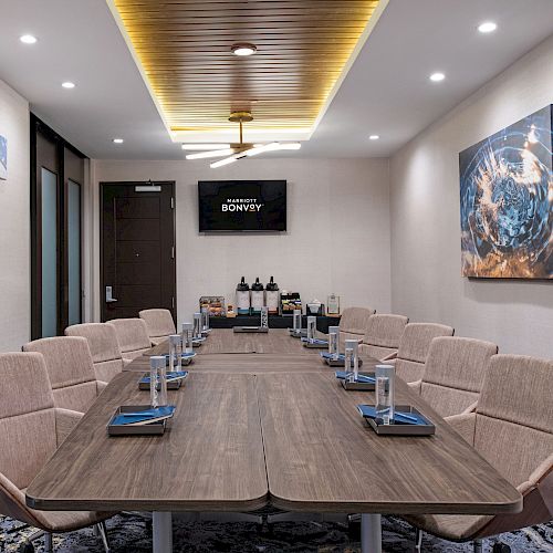 The image depicts a modern conference room with a long table, beige chairs, a TV, artwork on the walls, and a refreshment station at the back.