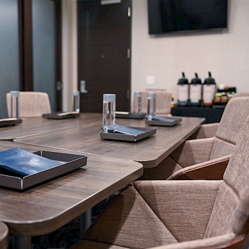 A conference room with a wooden table, six chairs, notebooks, and water bottles. A TV and a refreshment station are in the background, ending the sentence.