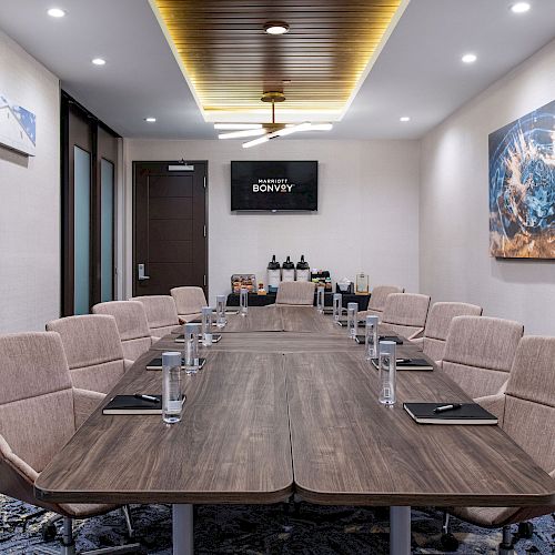 The image shows a modern conference room with a long table, twelve chairs, water glasses, notepads, wall art, a screen, and a door on one side.