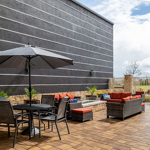 The image features a patio with outdoor furniture including a table with an umbrella, chairs, and cushioned seating. A cloudy sky is in the background.