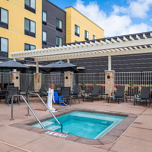 The image shows an outdoor area with a small rectangular pool, lounge chairs, tables with umbrellas, and a white pergola, next to a multi-story building.