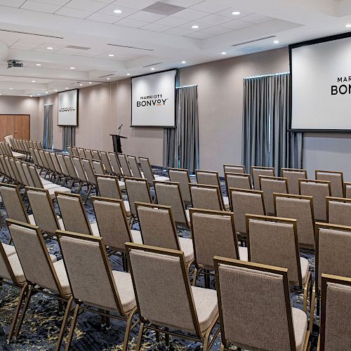 An empty conference room with rows of chairs facing two large screens displaying the 