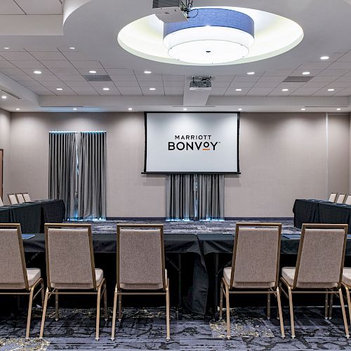 The image shows a conference room set up in a U-shaped seating arrangement with chairs and tables facing a screen displaying 