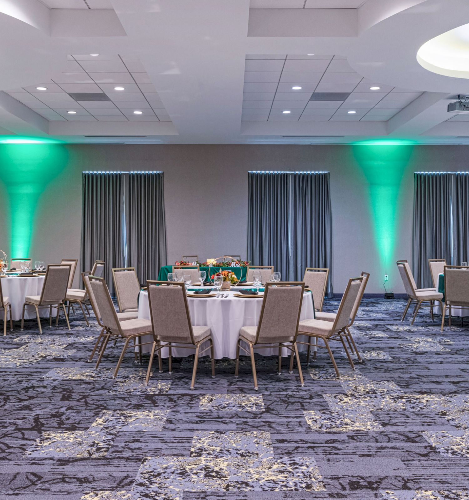 A banquet hall with round tables and chairs arranged, green accent lighting, and modern ceiling fixtures is shown in the image.