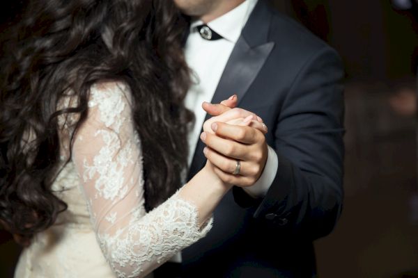 A couple is dancing closely, with the woman's long hair and lace dress, and the man wearing a suit, holding hands and embracing.