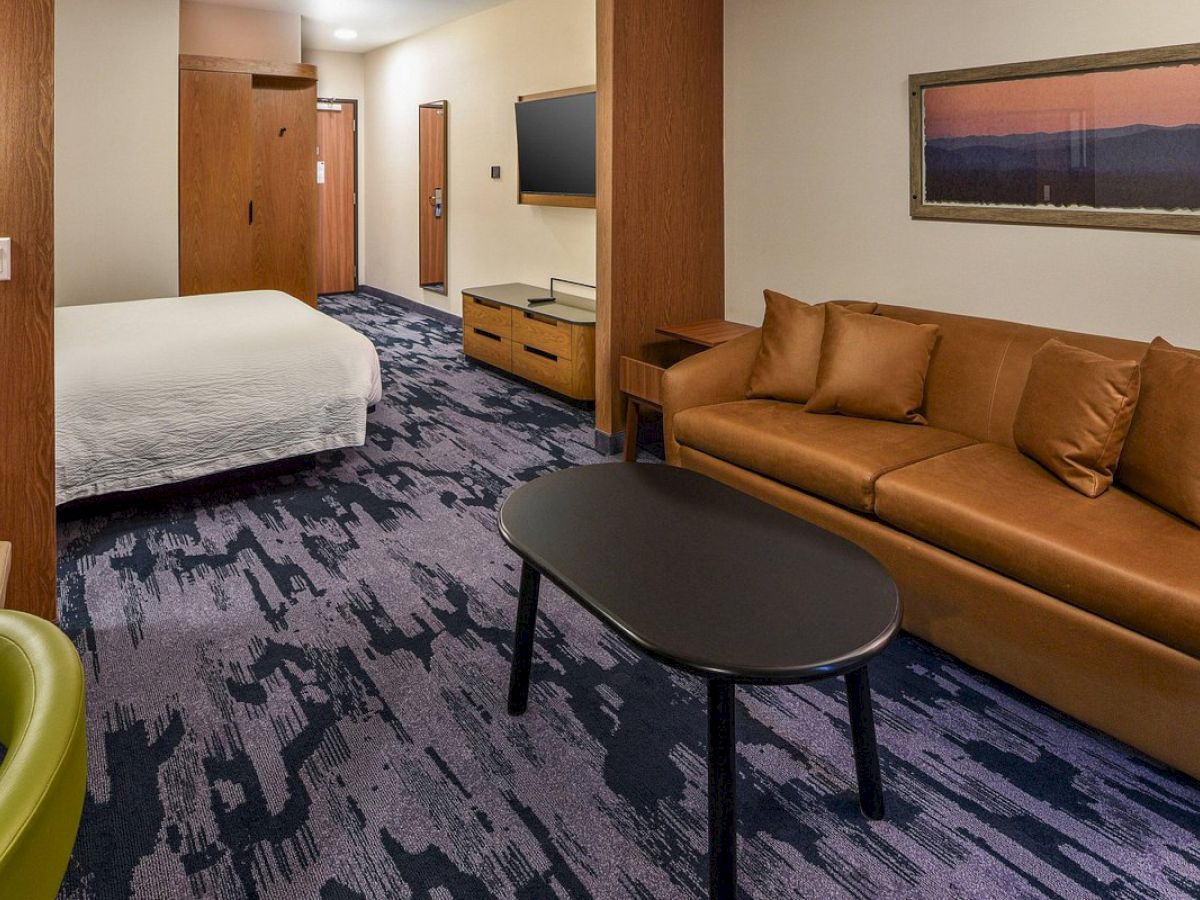 The image shows a hotel room with a bed, a couch, a coffee table, a TV, and a picture on the wall, featuring carpeted floor and modern decor.