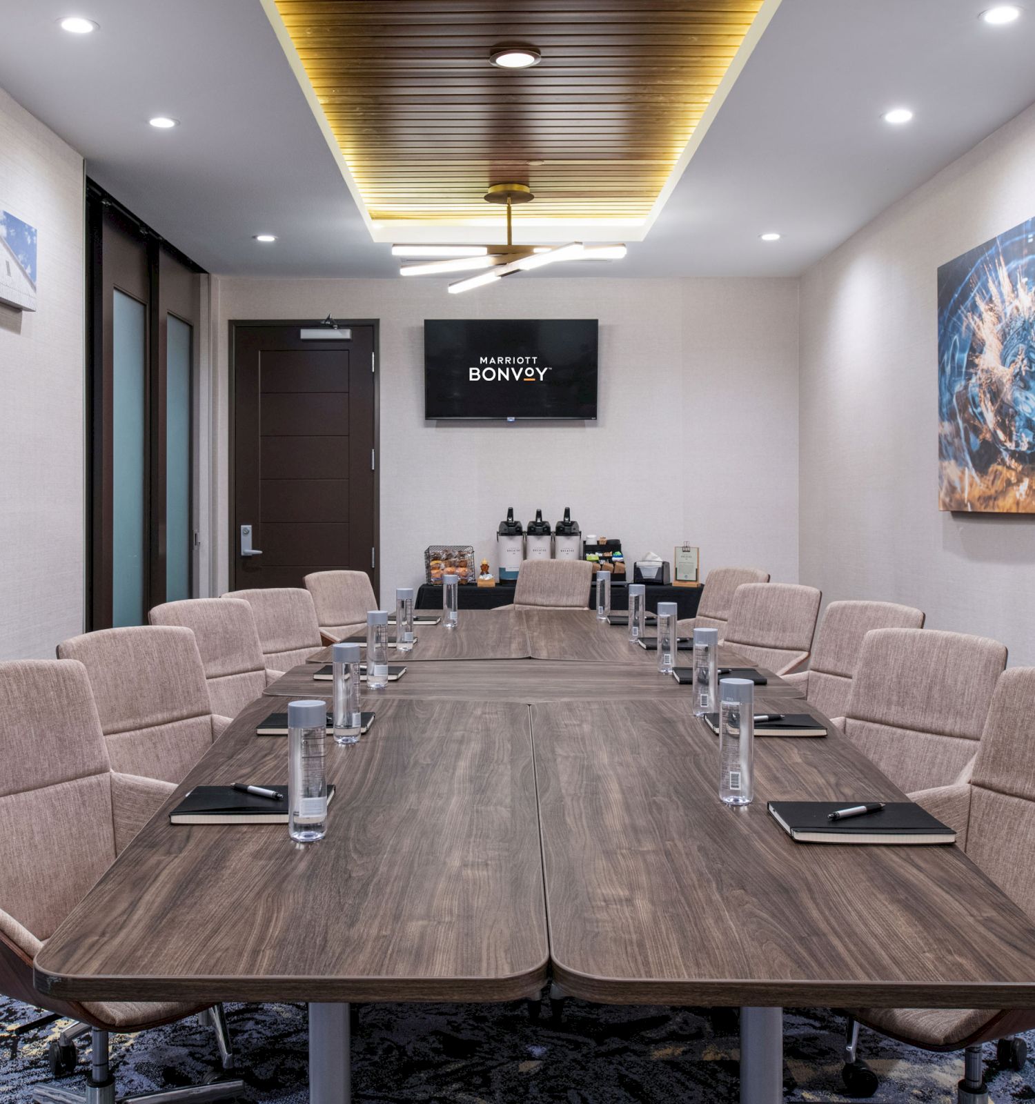 A modern conference room with a long table, chairs, notepads, water glasses, and a screen on the wall. There are artworks and a door in the background.