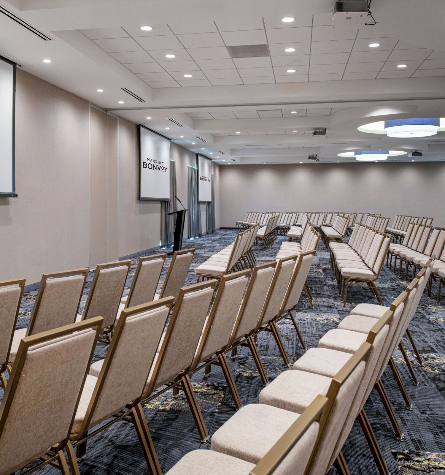 This image shows a conference room set up with rows of beige chairs facing two large screens displaying the Marriott Bonvoy logo. The room is well-lit.