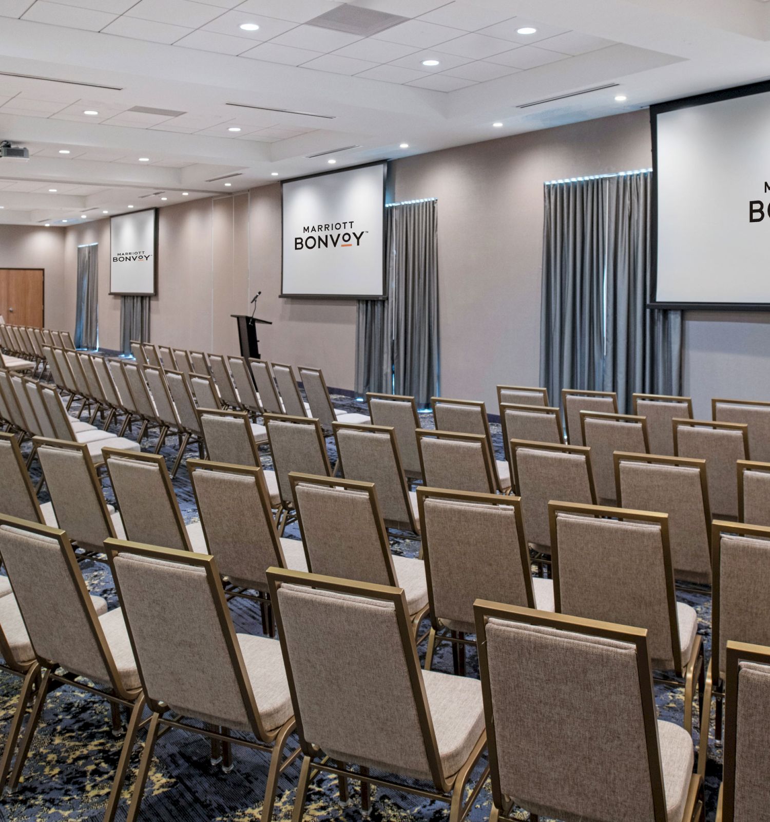 The image shows a conference room set up with rows of chairs, two screens displaying 