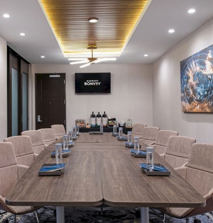 A modern conference room with a long table, beige chairs, a TV on the wall, and abstract artwork. It has a ceiling fan and a beverage station.