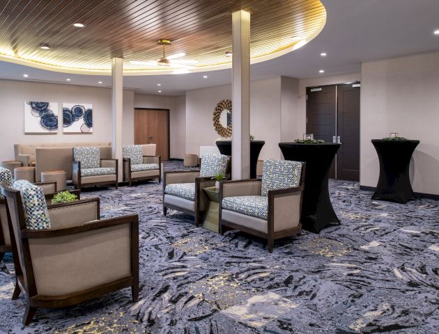 A modern lounge area with patterned armchairs, small tables with plants, artistic wall decor, a decorative mirror, and two tall black tables.