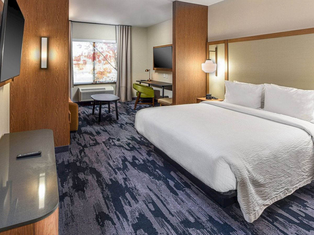 A modern hotel room with a large bed, TV, desk, chair, and a window with curtains. The decor features wooden accents and a patterned carpet.