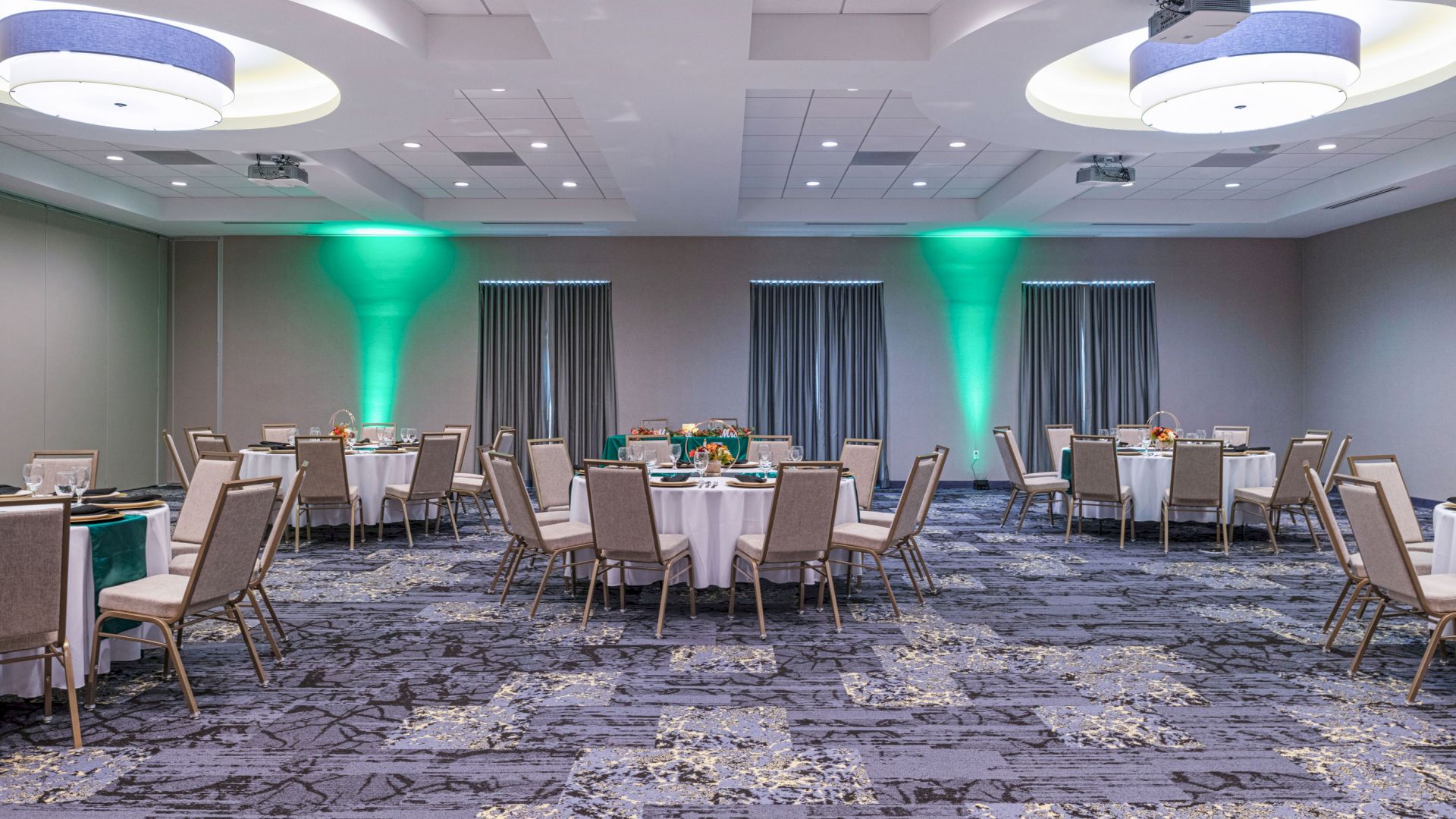 This image shows a decorated event room with round tables and chairs, subtle lighting, and green uplights on the walls.