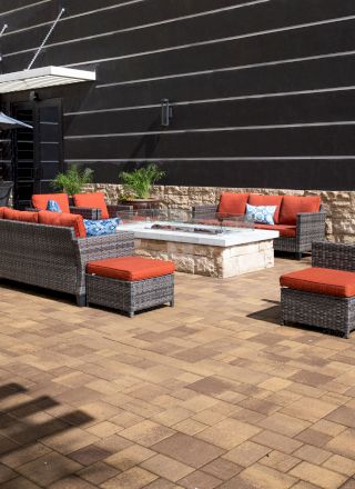 The image shows an outdoor patio area with wicker furniture, red cushions, a fire pit, potted plants, and a table with an umbrella.