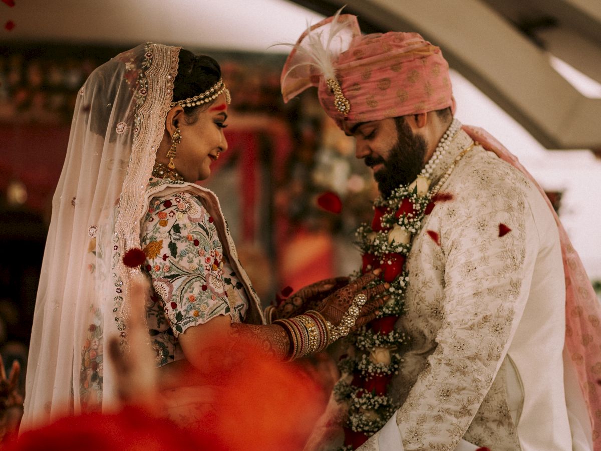 A couple dressed in traditional attire, exchanging wedding garlands during a ceremony with rose petals in the air.