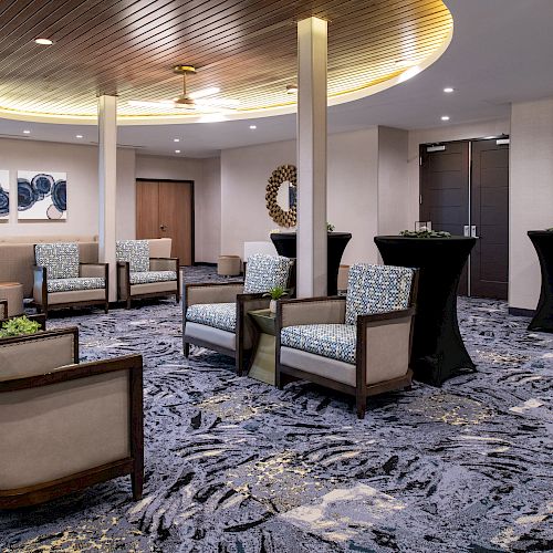 A modern lounge area with armchairs, round tables, wall art, a mirror, and doors, all set in a room with patterned carpet and a wooden ceiling.