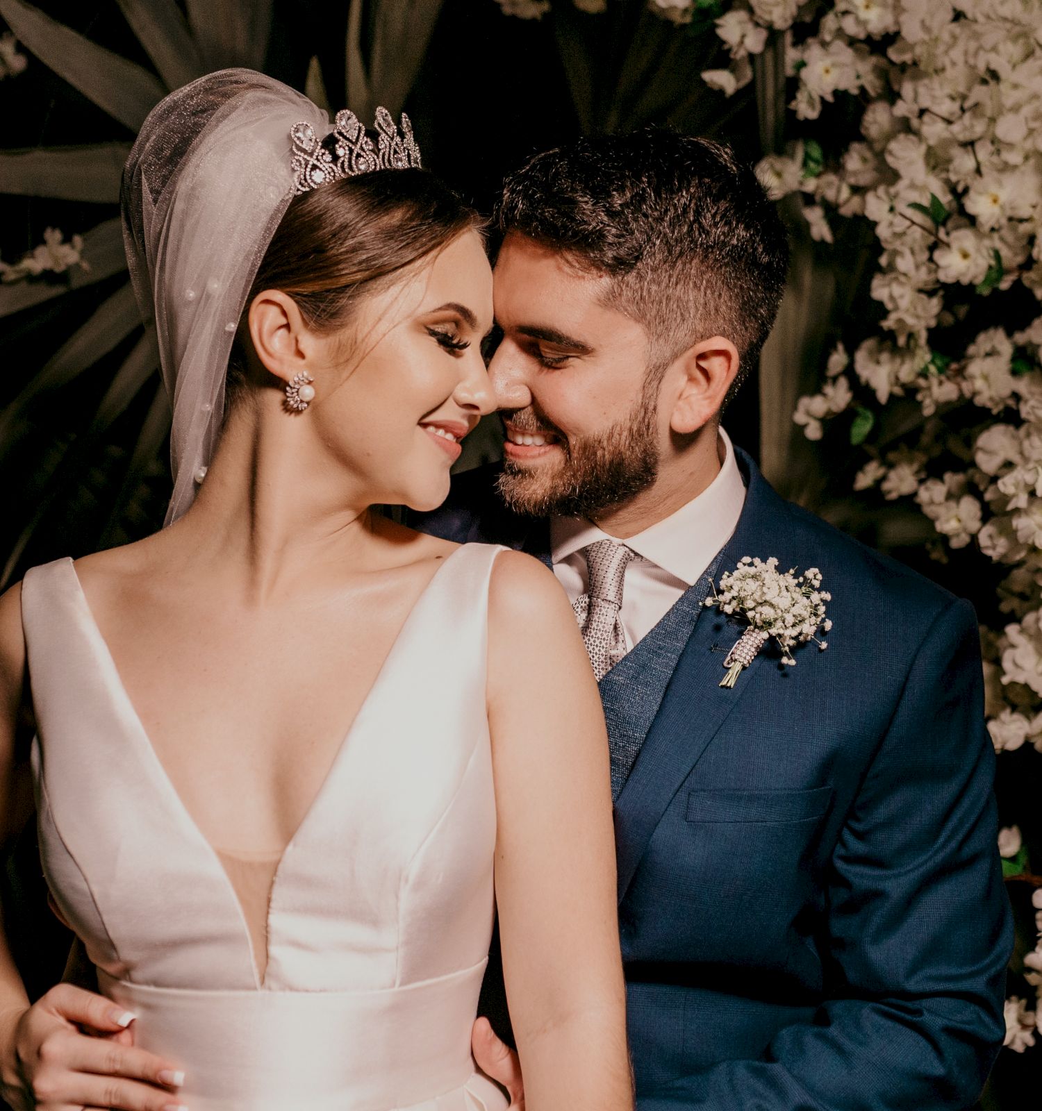 The image shows a bride and groom embracing, both smiling and looking at each other lovingly, standing in front of a flower-filled backdrop.