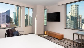 The image shows a bright room with large windows, a bed, a chair, a wall-mounted TV, and a bench, with a cityscape visible outside the windows.