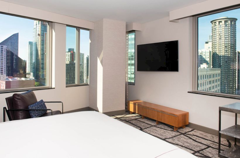 The image shows a bright room with large windows, a bed, a chair, a wall-mounted TV, and a bench, with a cityscape visible outside the windows.