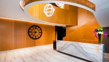 A modern lobby with a curved wooden ceiling, large wall clock, marble reception desk, and decorative light fixtures and flowers on the counter.